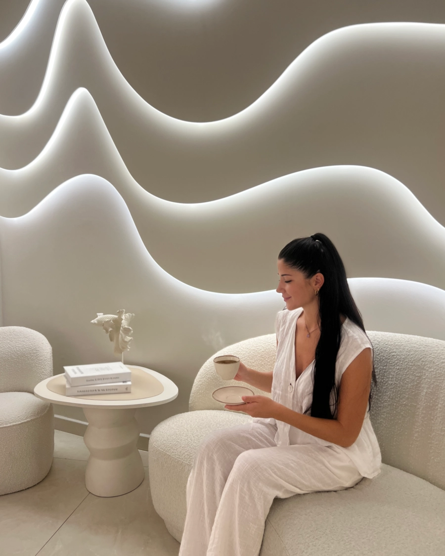 Una mujer disfruta de una bebida caliente en la sala de espera minimalista. La escena captura la experiencia completa de bienestar, desde la acogida hasta el relax absoluto antes o después de un tratamiento.